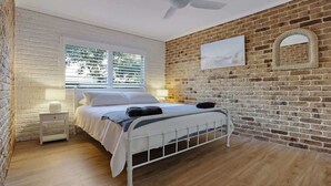 Una camera da letto con un letto, un comodino, una finestra con persiane, un muro in mattoni e una ventola a soffitto.