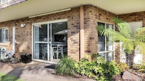 A brick building with a glass door entrance, a small garden with plants, and a visible patio area.