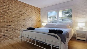Ein Schlafzimmer mit Backsteinwand, einem Bett mit weißer Bettwäsche, einem Fenster mit Jalousien und einem Nachttisch mit Lampe.