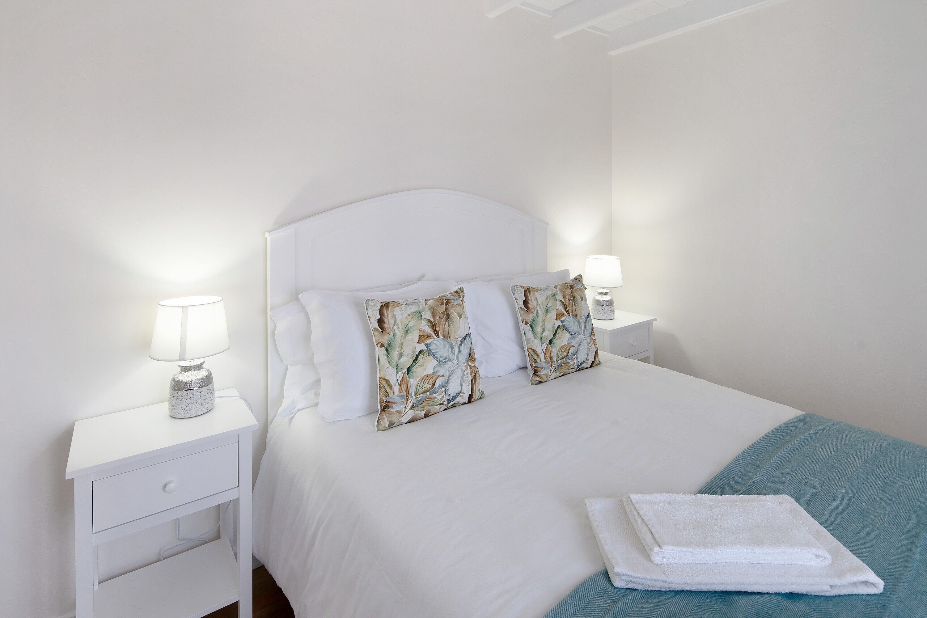 Un lit bien fait, avec une literie blanche et des coussins à motifs floraux, une table de chevet blanche avec une lampe, et une serviette blanche pliée posée sur le lit.