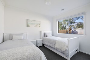 Two single beds with white bedding and headboards, a bedside table and lamp, and a large window with a view of the outdoors.