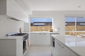 Une cuisine moderne avec des meubles blancs, une cuisinière à gaz et une grande fenêtre donnant sur une clôture en bois.