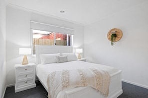 Un dormitorio blanco con cama, dos mesitas de noche, una ventana y un estante circular fijado a la pared.