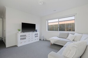 Un salon moderne avec un téléviseur à écran plat posé sur un meuble blanc, un canapé gris et une grande fenêtre donnant sur un mur de briques.