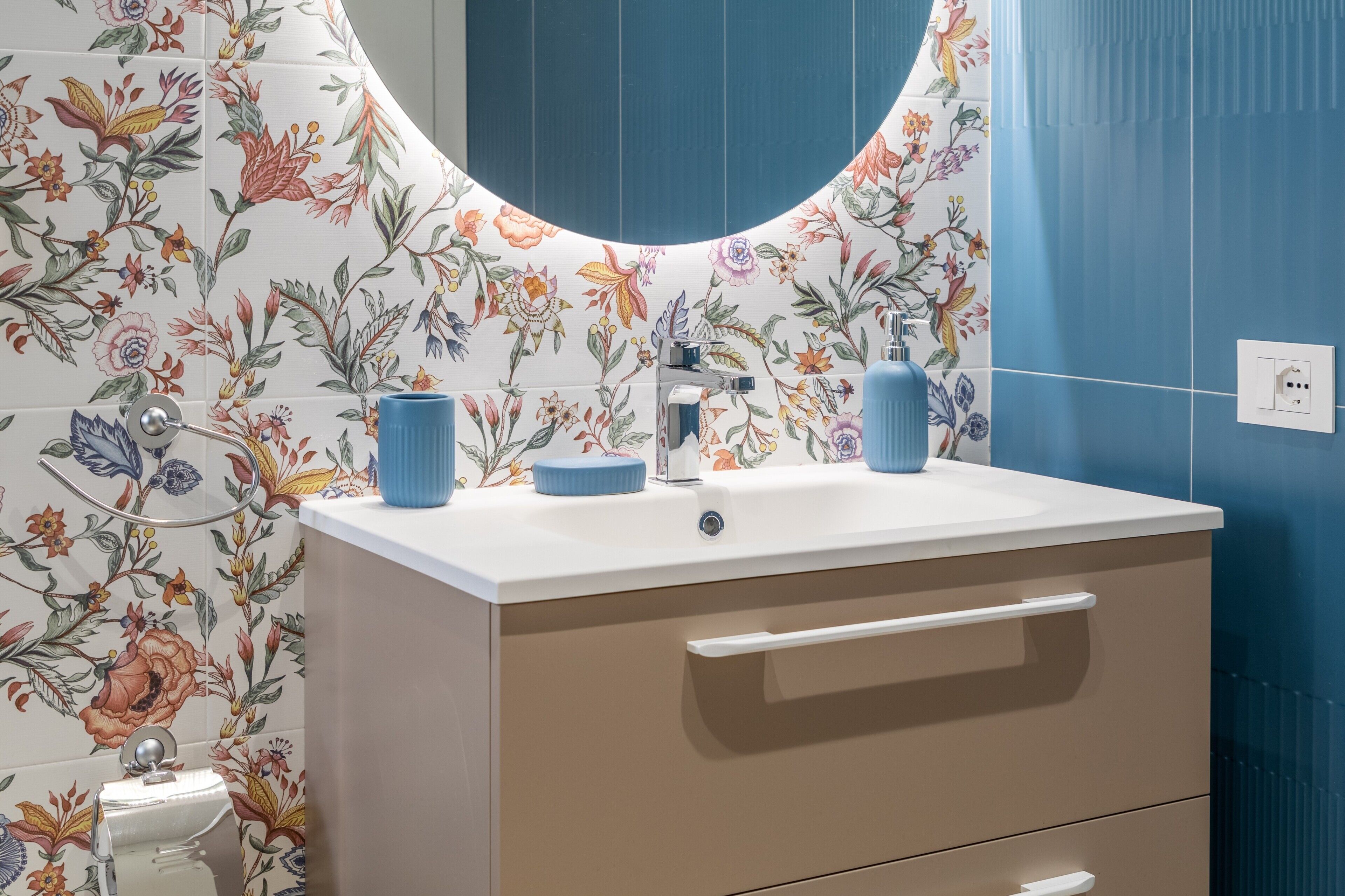 Un baño con salpicadero de azulejos con motivos florales, un lavabo blanco, un grifo azul y un espejo.