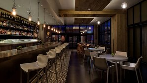 Een bar met een lange toonbank, meerdere stoelen en diverse flessen en glazen op planken.