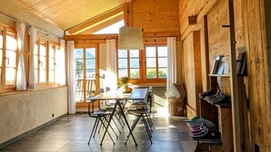 A spacious room with a high wooden ceiling, a dining table with chairs, a large window, and a shoe rack. - Apartment Chalet Grittelihus, between Interlaken - Gstaad (Diemtigen)