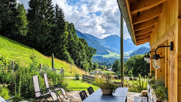 A stone patio with a table and chairs, a wooden pergola, and a view of a green landscape with trees and mountains. - Apartment Chalet Grittelihus, between Interlaken - Gstaad (Diemtigen)