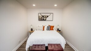 Un dormitorio con una cama grande, dos mesitas de noche con lámparas, un cuadro enmarcado en la pared y dos taburetes tapizados.