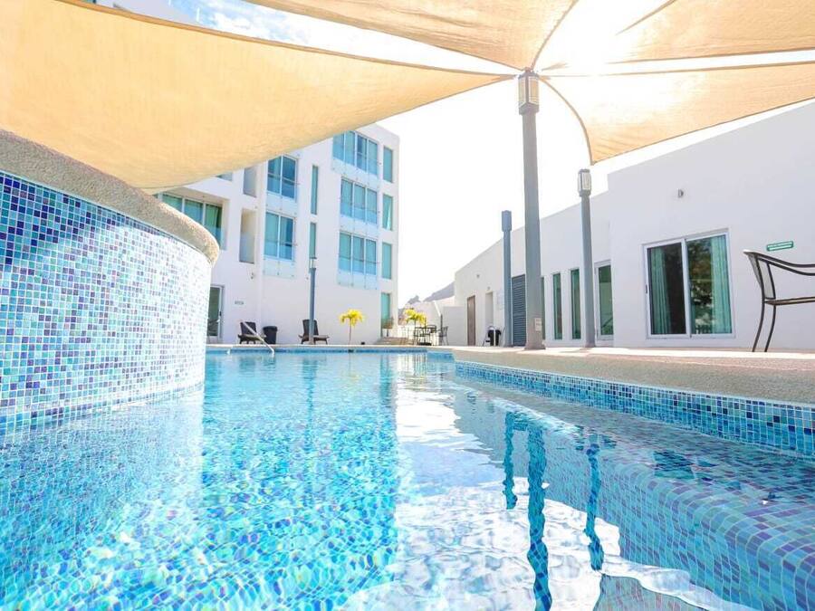 Una piscina con mosaicos azules, rodeada por un edificio blanco con ventanas amplias y una zona de estar cubierta.