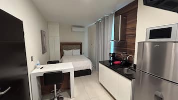 Habitación de hotel compacta con cama, escritorio con silla, kitchenette con refrigerador y una ventana pequeña con cortinas.