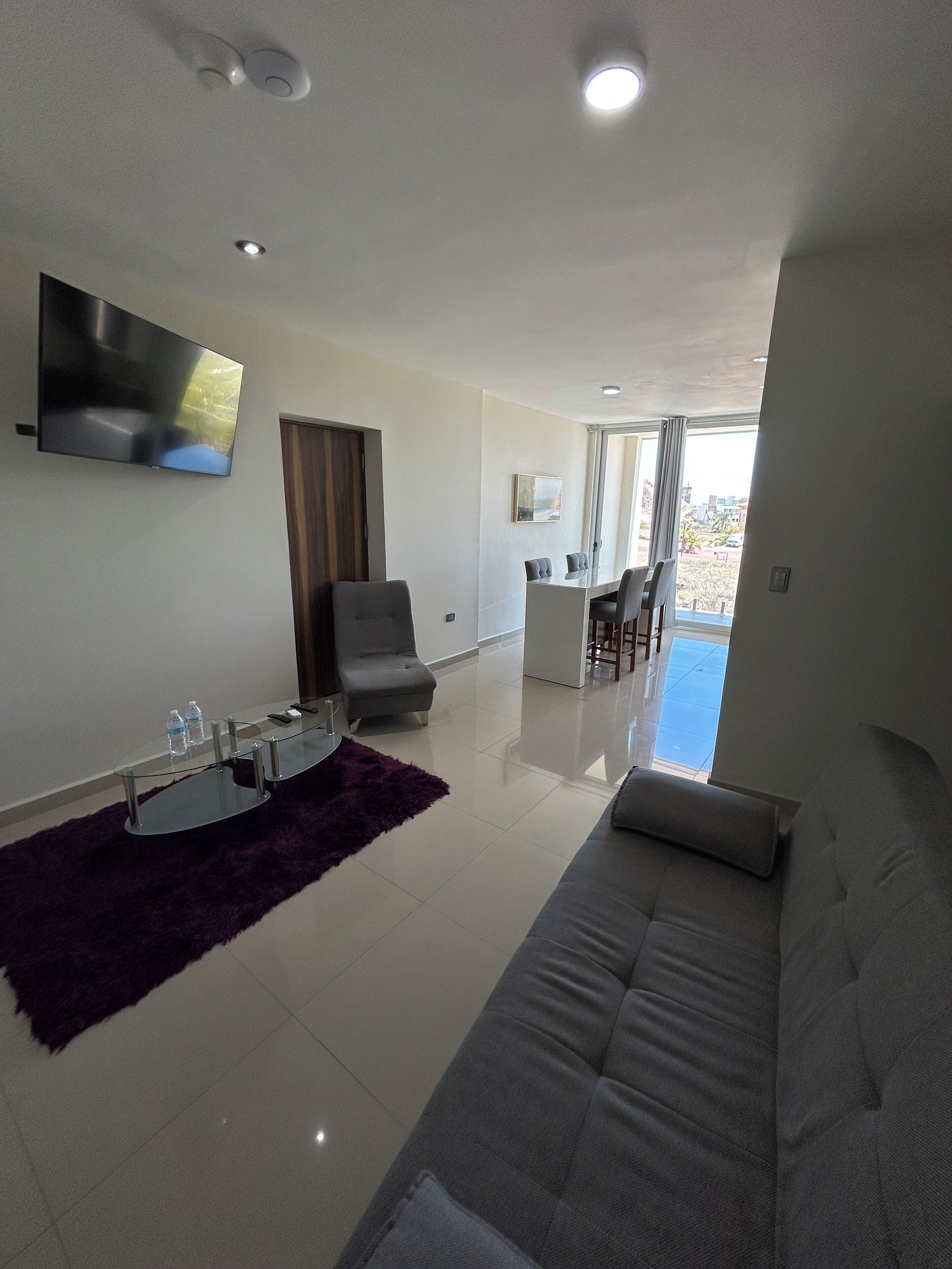 A modern living room with a sofa, a glass coffee table, a flat-screen TV, and a dining area with chairs and a table.