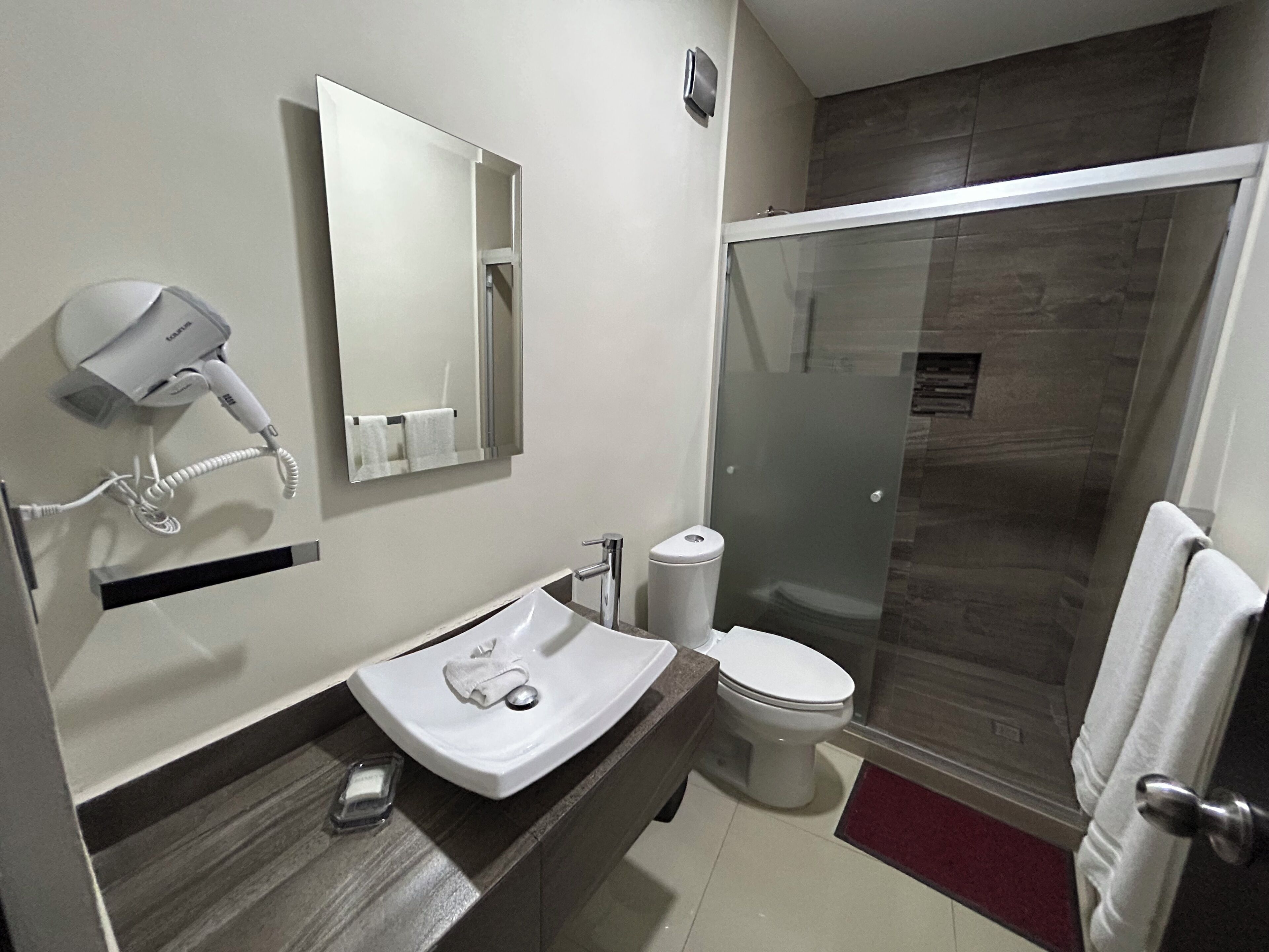 Un baño moderno con lavamanos blanco, secador de pelo, espejo, inodoro y cabina de ducha de vidrio.