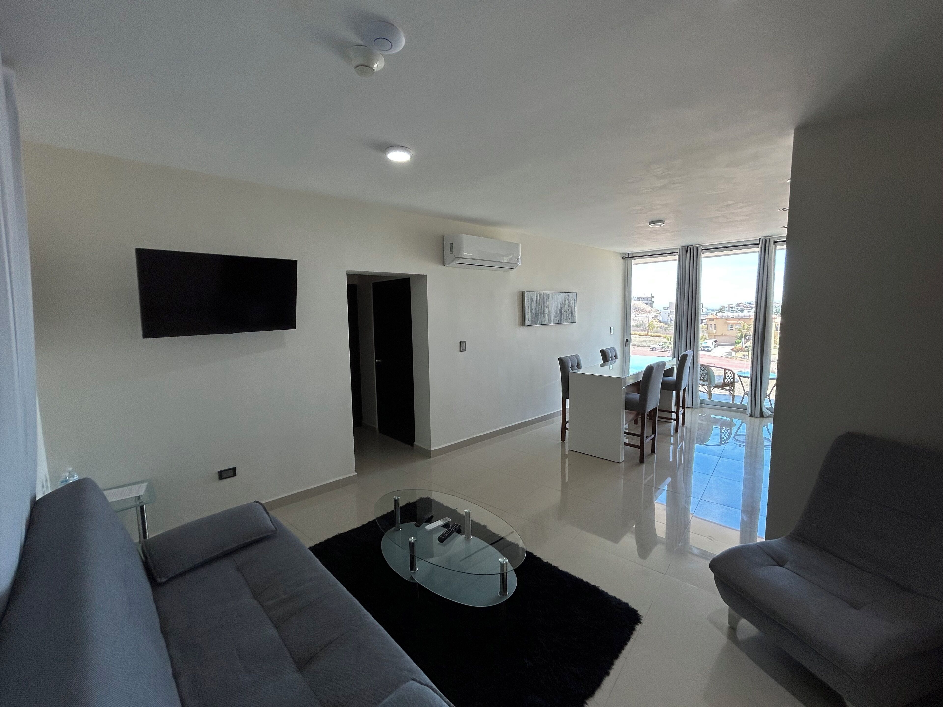 A modern living room with a sofa, a coffee table, and a TV mounted on the wall. There is a balcony with a view of the city.