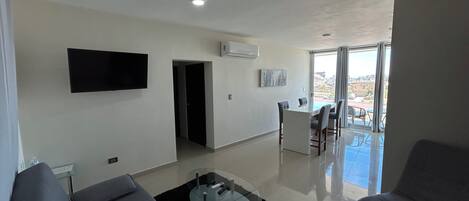 Una sala de estar moderna con un sofá, una mesita de centro y un televisor montado en la pared. Hay un balcón con vistas a la ciudad.