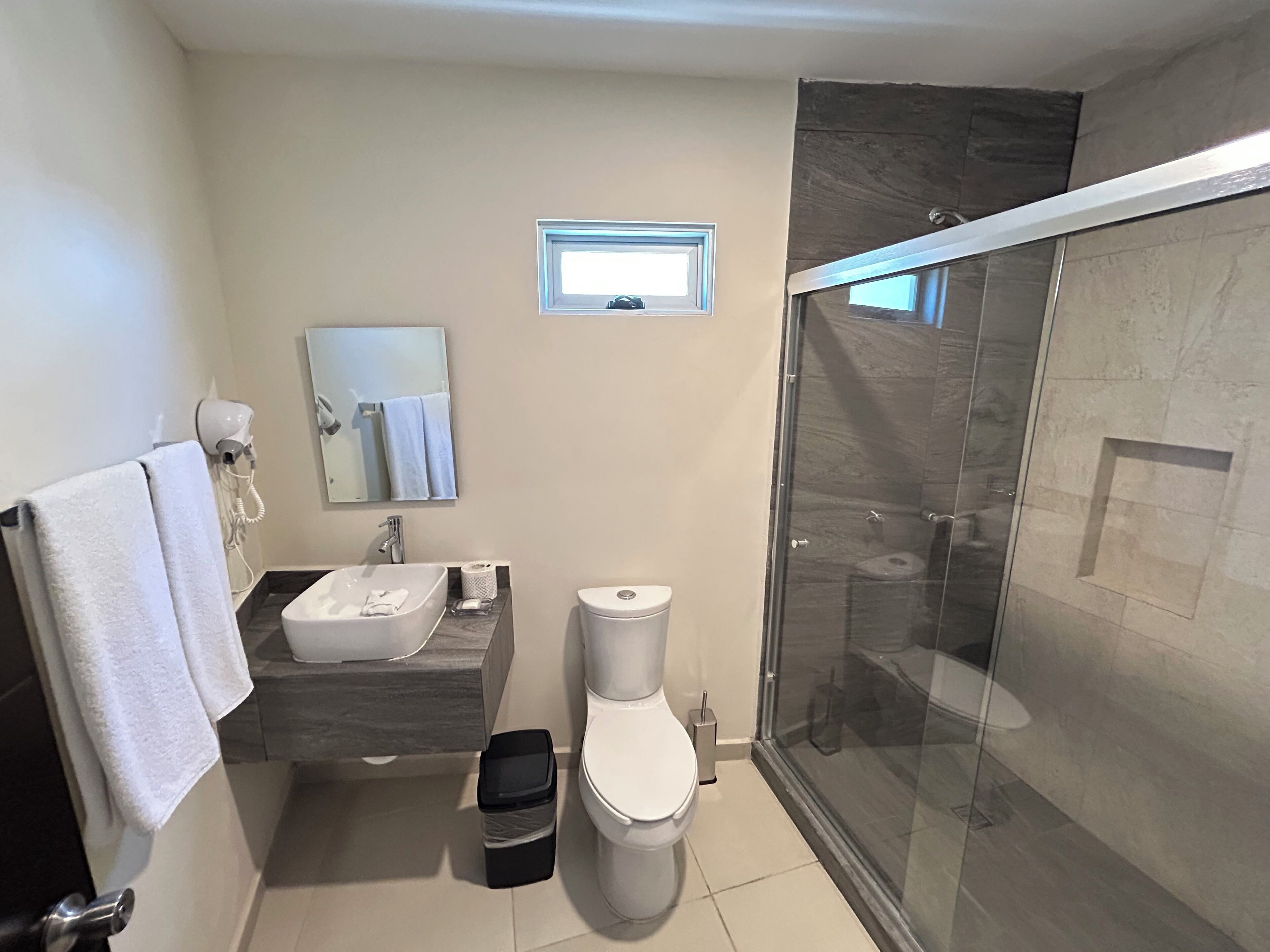 Une salle de bain moderne avec une douche à parois de verre, un lavabo blanc, des toilettes et une petite fenêtre.