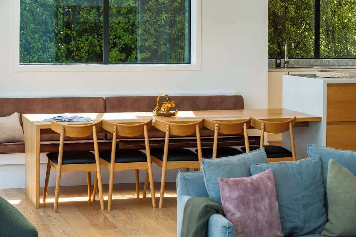 Een moderne woonkamer met een houten eettafel, vier stoelen, een bank en een groot raam met uitzicht op groen.