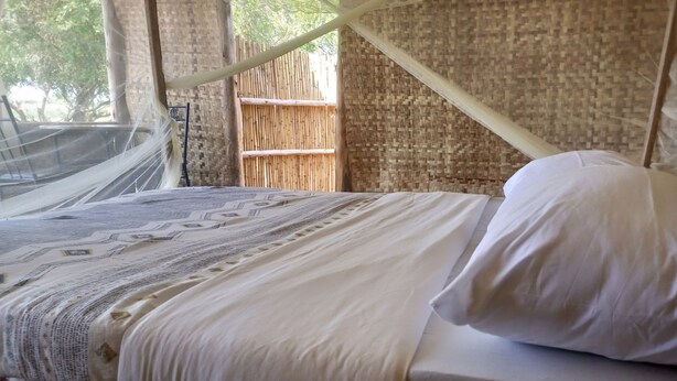 A bed with white linens and a canopy, set against a bamboo wall and a woven mat.