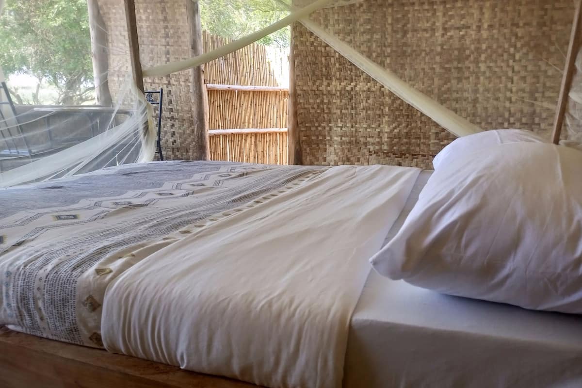A bed with white linens and a canopy, set against a bamboo wall and a woven mat.
