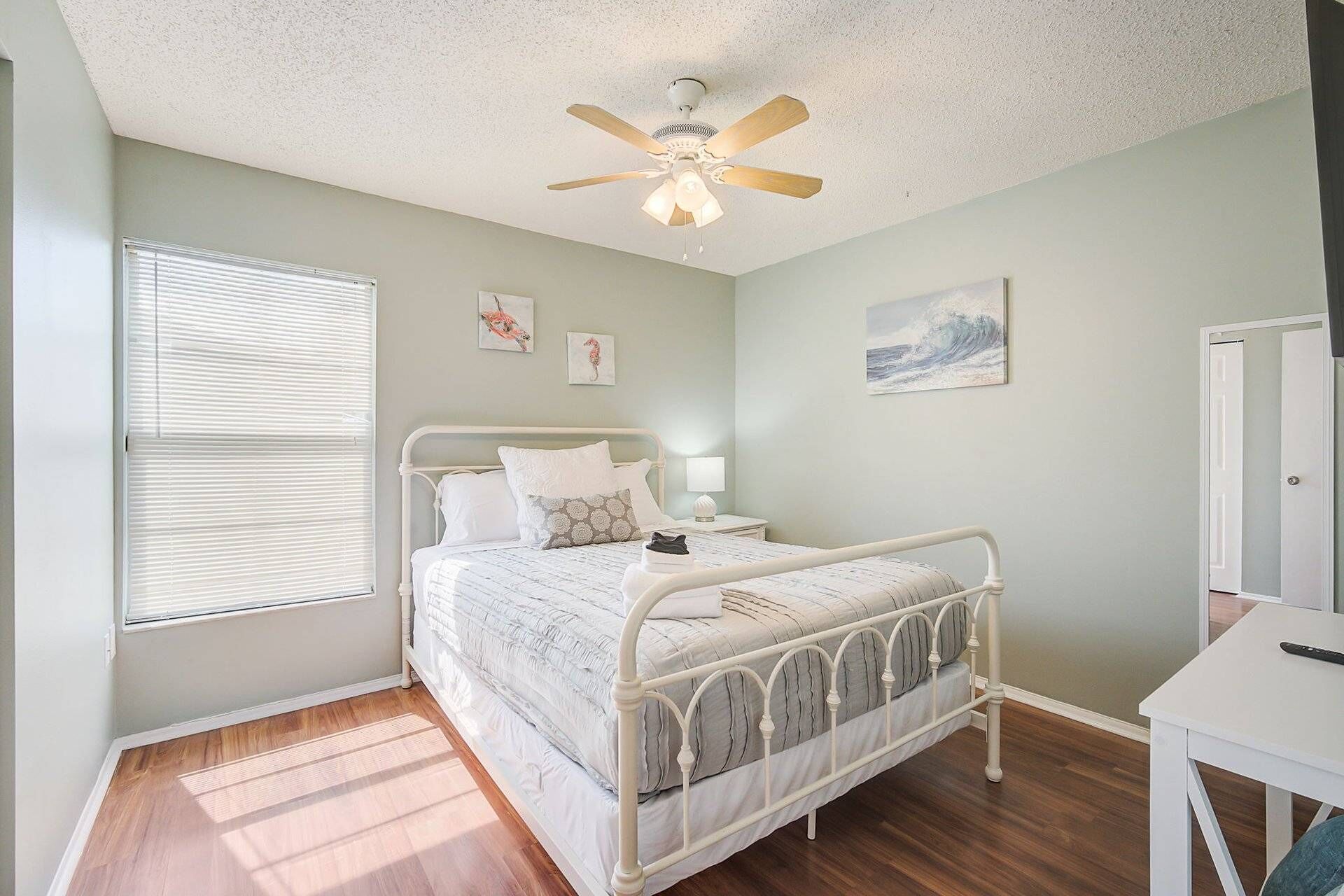 A bedroom with a bed, a ceiling fan, a window with blinds, and artwork on the walls.