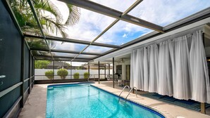 An indoor swimming pool with clear blue water, surrounded by a glass enclosure, and a covered patio area with white curtains. - Family Oasis--Private Pool & Hot Tub-Near Anna Maria Island, Beaches, Parks, Shops (Bradenton)