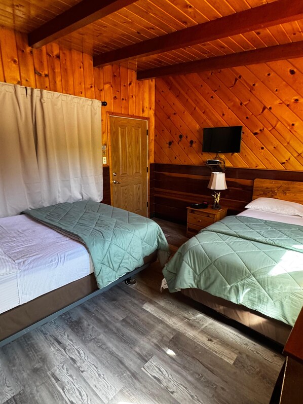 A room with two beds, wooden walls, a door, a nightstand with a lamp, and a TV on the wall. - Toiyabe Motel (Coleville)