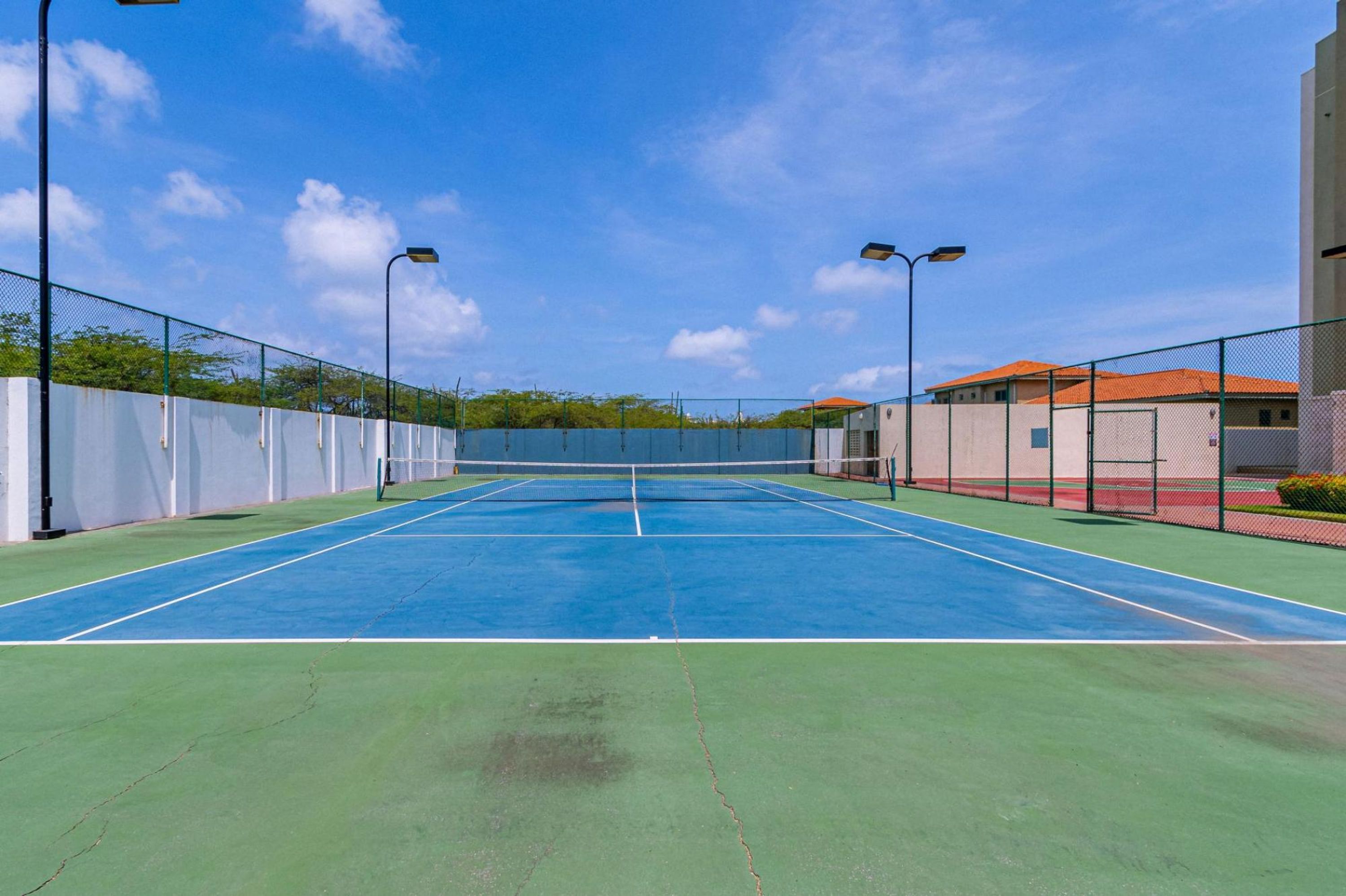 Een tennisbaan buiten, met een net, omheind door een hek en straatverlichting.
