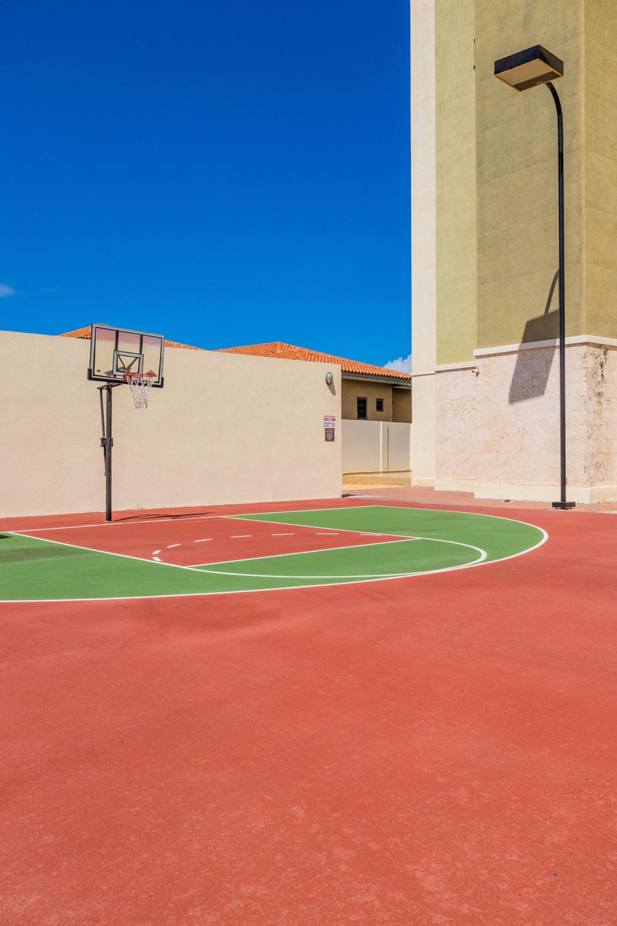 Ein Basketballplatz im Freien mit roter Spielfläche und einer Grünanlage, umgeben von einer beigen Mauer und einem hohen Laternenmast.