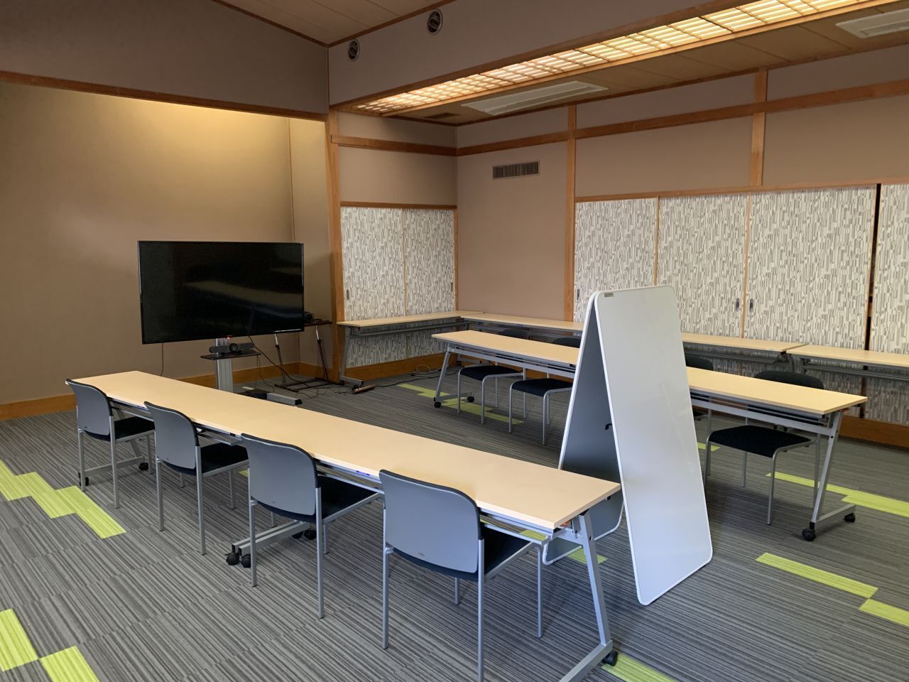 Een vergaderruimte met lange tafels en stoelen, een flatscreen-tv en een whiteboard.