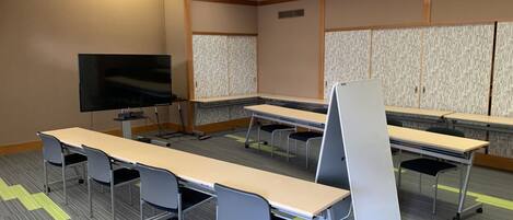 Un salón de conferencias con mesas largas y sillas, un televisor de pantalla plana y una pizarra.