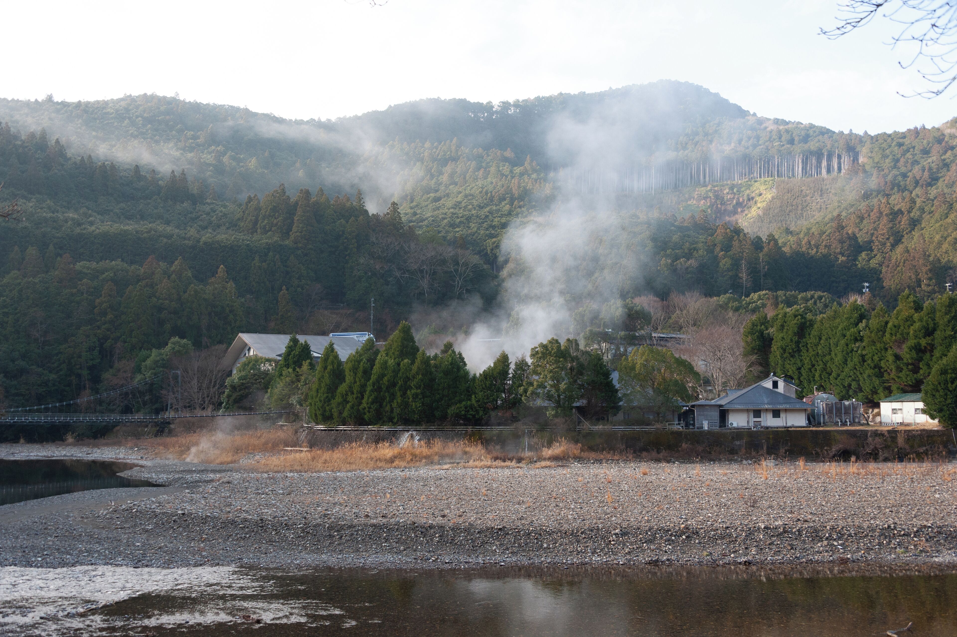 A riverside view with a building, trees, and hills in the background.