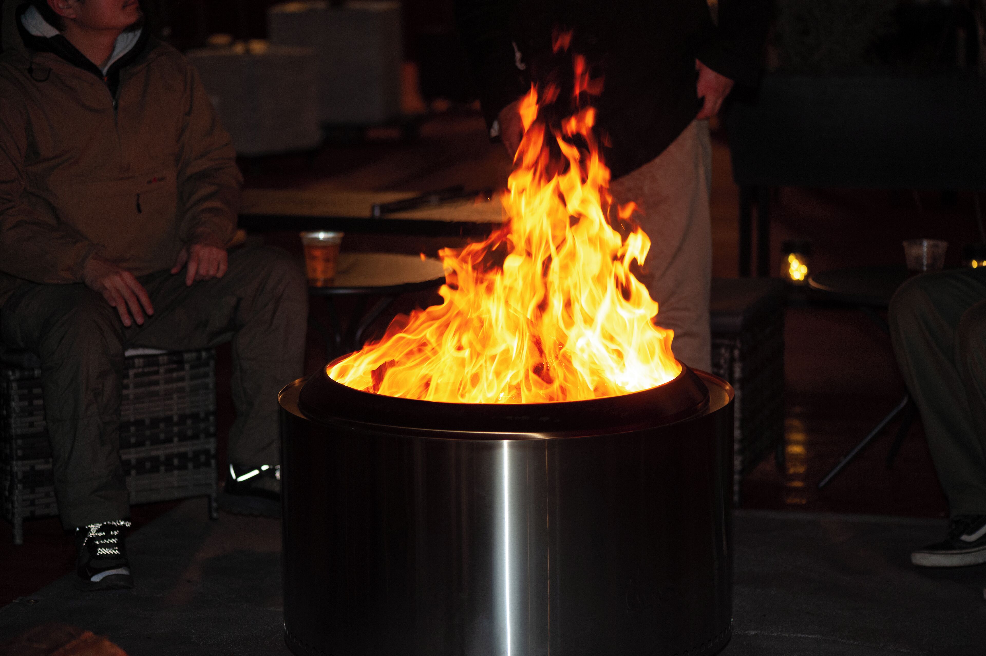 A metal fire pit with flames, surrounded by people sitting on chairs.