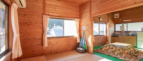 Interior de una cabaña de madera con suelo de tatami, un futón, una mesita y una zona de cocina visible a través de una puerta abierta.