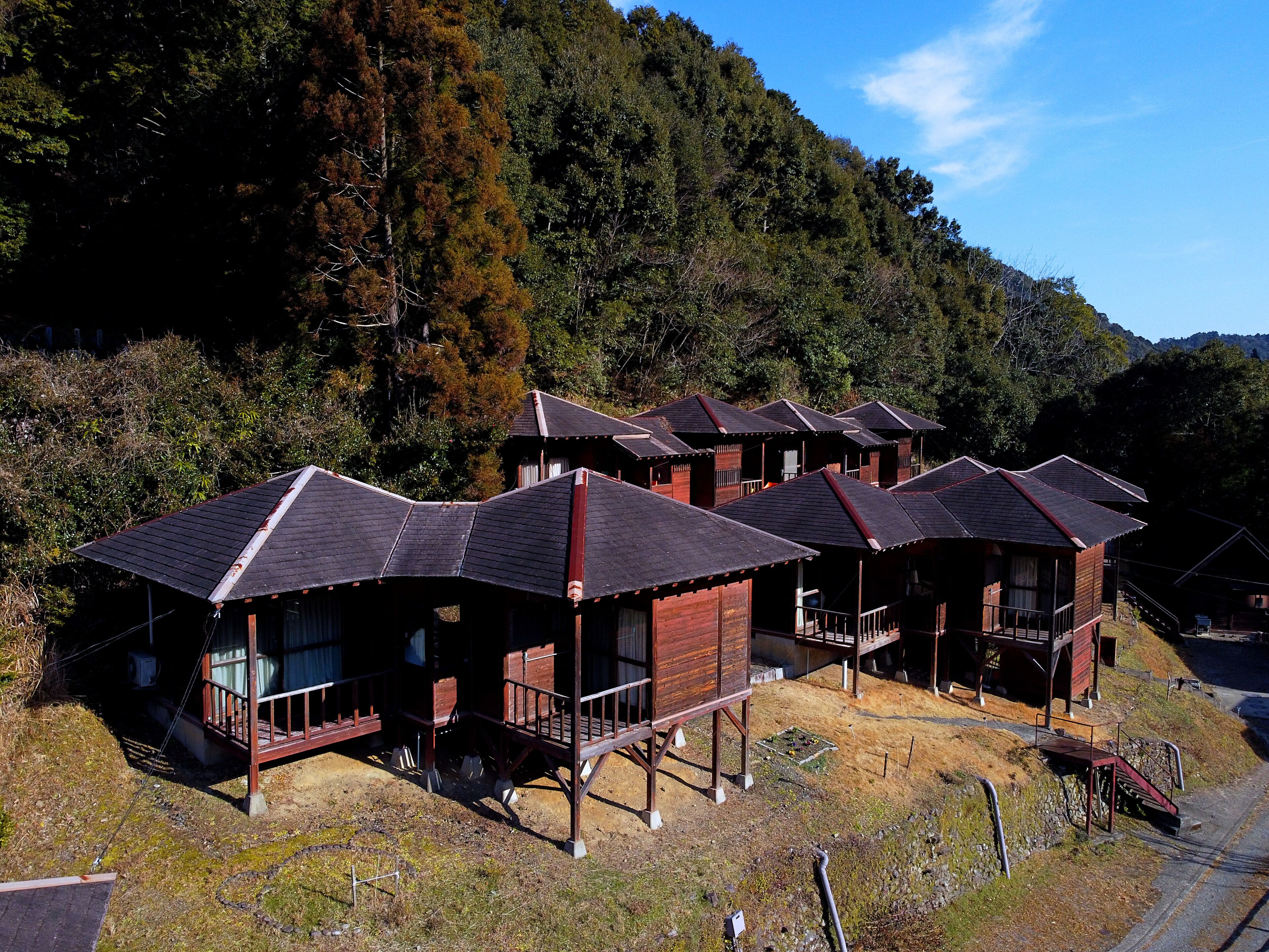 A row of wooden cabins with sloped roofs, set against a backdrop of dense forest and hills.