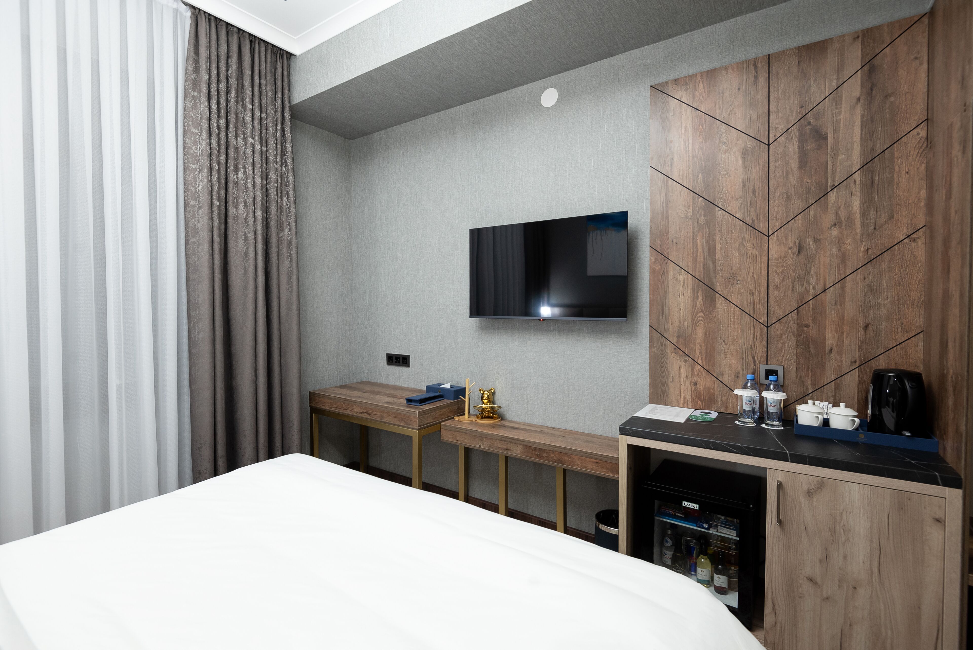 A modern hotel room with a bed, a TV mounted on the wall, a wooden desk, and a minibar.