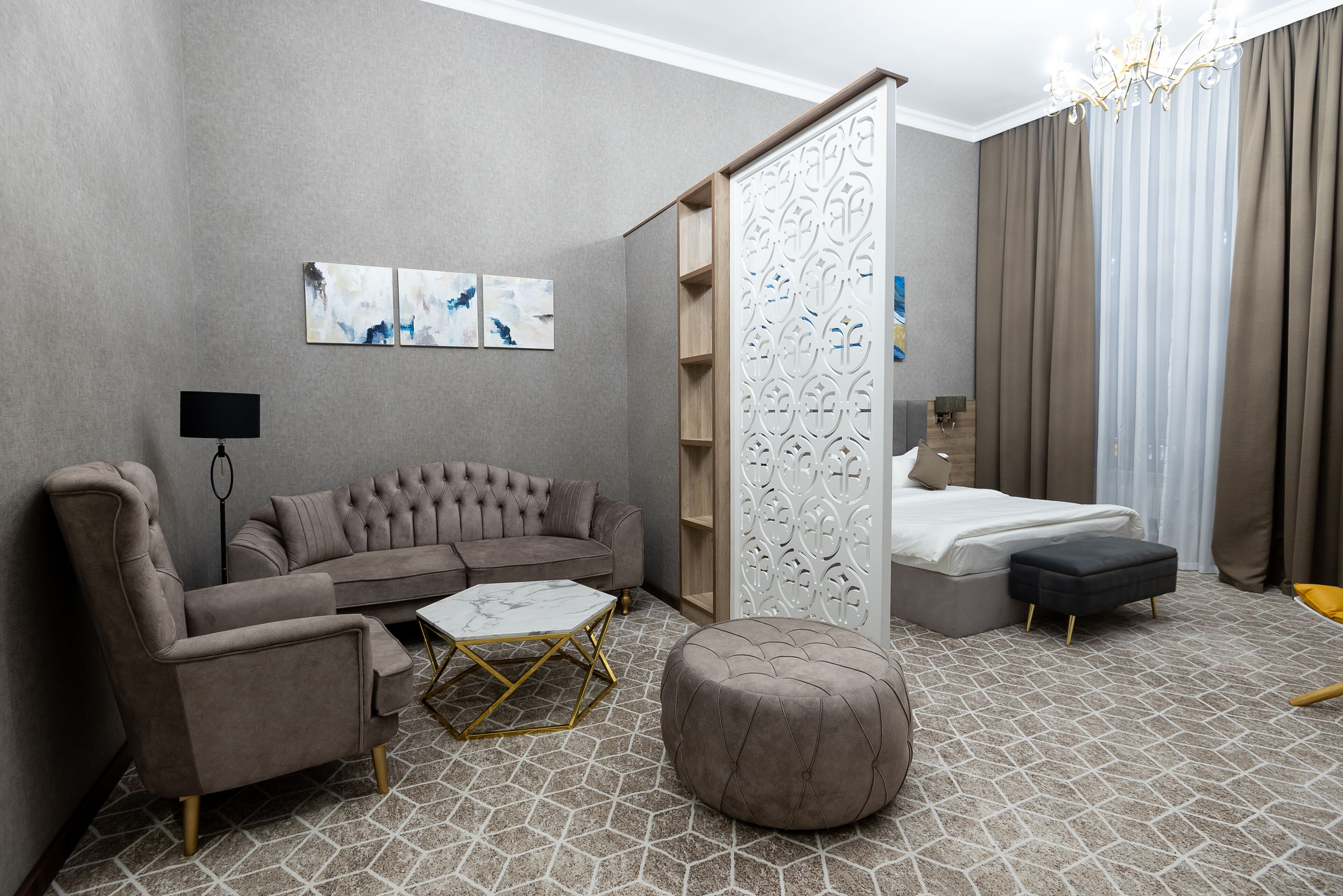 A modern hotel room with a grey sofa, a round ottoman, a white coffee table, and a bed with a grey headboard.