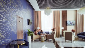 Un moderno vestíbulo de hotel con un diseño de pared azul y dorado, una gran lámpara de araña y un televisor de pantalla plana montado en la pared.