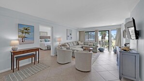 A spacious living room with a large sectional sofa, a coffee table, and a TV mounted on the wall. There is a dining area with a table and chairs, and a balcony with a view of palm trees.