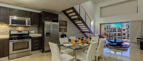A modern kitchen with stainless steel appliances, a dining area with white chairs, and a staircase leading to an upper level.