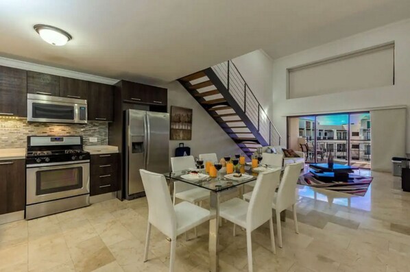 A modern kitchen with stainless steel appliances, a dining area with white chairs, and a staircase leading to an upper level.