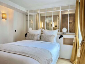 A modern hotel room with a large bed, a bedside table with a lamp, a mirror, and a window with curtains. - Elegant family flat with lift - Luxembourg Gardens (Paris)