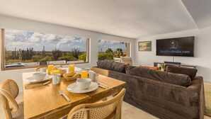 A modern living room with a dining area, large windows with desert views, a flat-screen TV, and a sign for VacationAruba.com. - Breathtaking Views 2br3ba Private Pool Large Space (Noord)