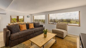 A modern living room with a dark brown sofa, a beige armchair, and a wooden coffee table. There are large windows offering views of a desert landscape. - Breathtaking Views 2br3ba Private Pool Large Space (Noord)