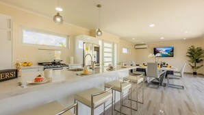 A modern kitchen with a central island, bar stools, and a dining area with a TV and plant. - NEW 3BR 3BA Villa w Modern Decor and Private Pool (Noord)