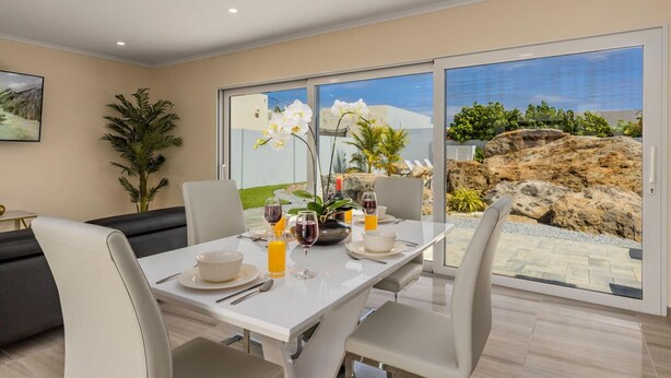 A modern dining area with a table set for two, featuring a view of a rocky landscape and greenery through large glass doors. - NEW 3BR 3BA Villa w Modern Decor and Private Pool (Noord)