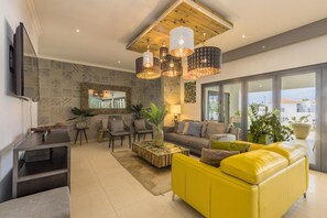 Ein modernes Wohnzimmer mit einem gelben Sofa, einem Couchtisch und einem Fernseher. Mehrere Stühle und ein großer Spiegel sind an der Wand angebracht.