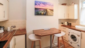 Cocina compacta con encimera de madera, un taburete, lavadora y un cuadro con un atardecer sobre un puerto.