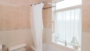 Un baño con bañera, inodoro, ventana con cortina y un pequeño barco modelo en el alféizar.