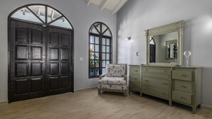 Ein Zimmer mit einer großen, dunkelholznen Tür, einem Fenster mit Ausblick, einem grünen Polstersessel und einem Schminktisch mit Spiegel.