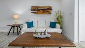 A modern living room with a white sofa, blue cushions, a wooden coffee table, a lamp, and a wall-mounted wooden art piece. - NEW 2BR Condo in of Palm Beach Walk 2 Beach (Noord)